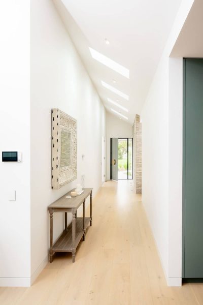 A light-filled hallway by luxury custom home builders in Melbourne featuring pale wood floorboards and a run of skylights.