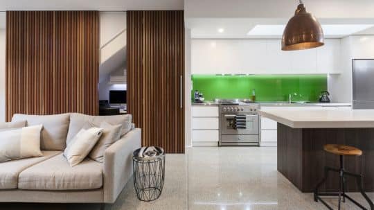 A home renovation displays a living space and kitchen with neutral tones and a refreshing green kitchen splashback.